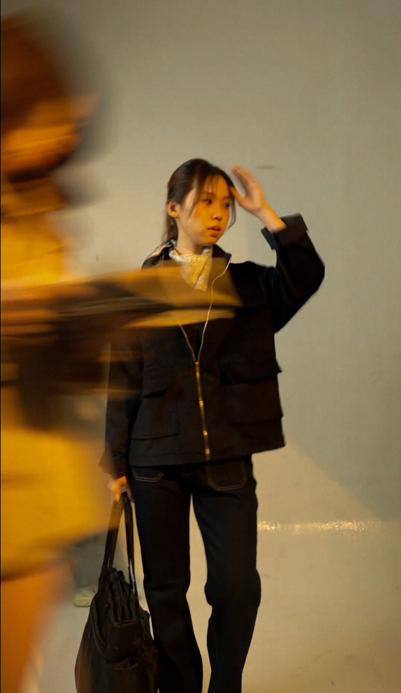 Model in the navy longneck jacket with a brass zip, hand raised, as someone blurs past