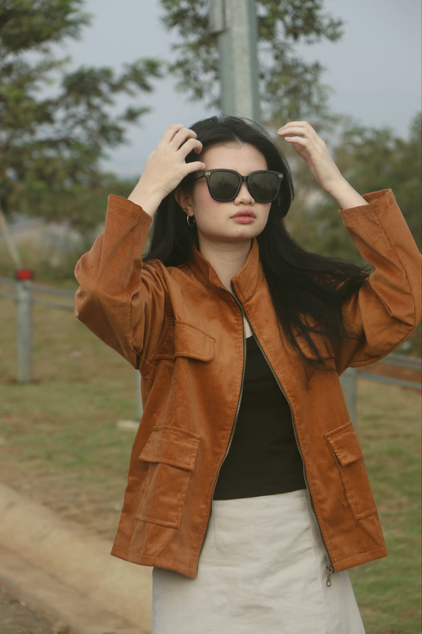 Model in sunglasses and the caramel longneck jacket, hands raised to her hair